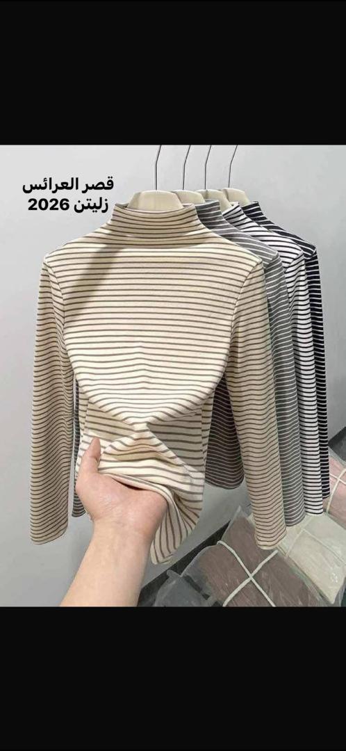 بلايز تيرتل مخططة بأكمام طويلة معلقة على شماعات مع يد تمسك بطرف القميص الأمامي لإظهار ملمس القماش، تدرج الألوان من البيج إلى الرمادي والأسود على خلفية محايدة داخل متجر عرض، يظهر نص في الصورة قصر العرائس زليتن 2026، الطابع أنيق وراقي