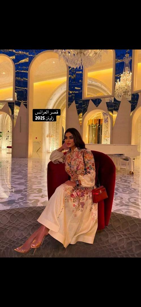 امرأة ترتدي طقماً أنيقاً مكوناً من بلوزة وسروال، تقف في متجر ملابس مع خلفية تعرض مجموعة متنوعة من الأزياء. النص الموجود في الصورة يقرأ قصر العرائس زليتن 2026، مما يضفي طابعاً عصريًا وثقة على المشهد.